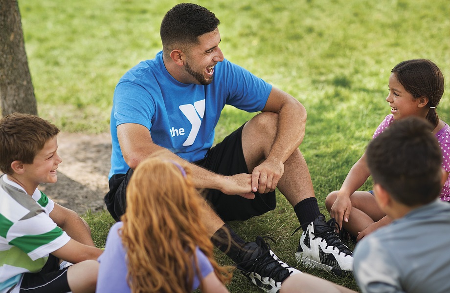 YMCA counselor in blue YMCA logo t-shirt sitting in the grass talking to campers