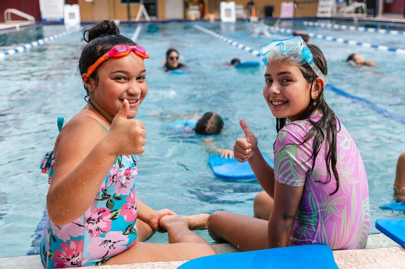 Two girls sitting on the edge of the pool wearing swimsuits and goggles giving the thumbs up at the Winter Park YMCA