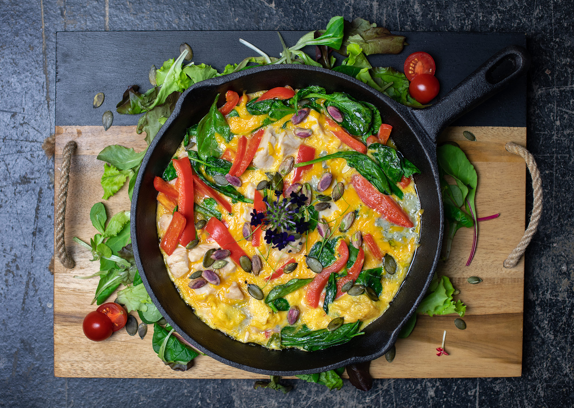 Healthy frittata with spinach, tomatoes, and other vegetables in cast iron pan on top of a cutting board
