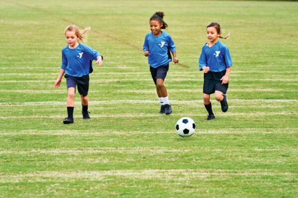 Three girls running after soccer ball on soccer field wearing YMCA logo uniforms