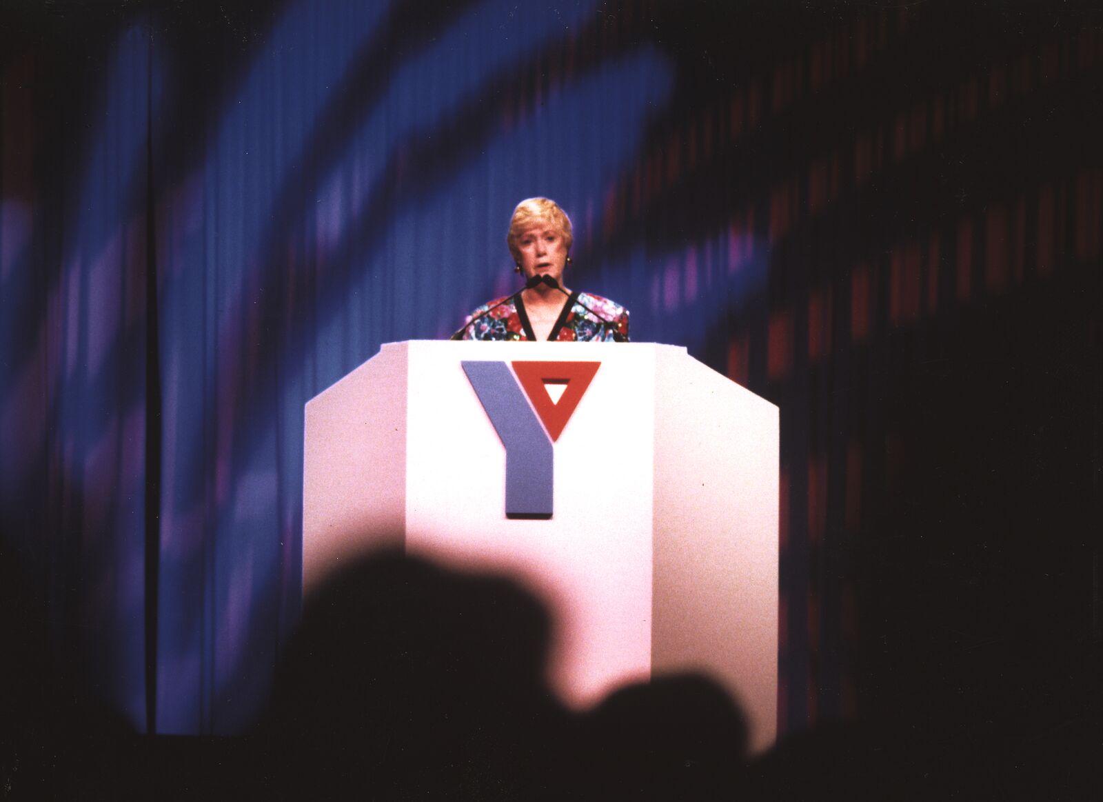 Image of Barbara Roper standing in front of blue curtains at a white podium with the old Y logo on it