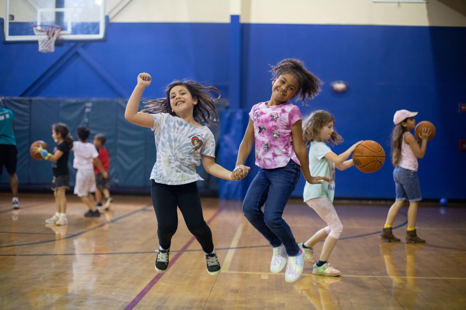 Two elementary aged girls jumping in a gym surrounded by playing children