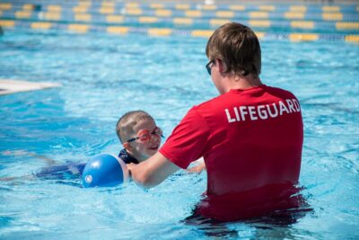 Lifeguard wearing red lifeguard t-shirt holding a pool floaty while a child swims and smiles