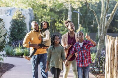 An African-American family with three children visiting a park, walking along a pedestrian walkway surrounded by lush foliate. The boy is 10 years old and the girls are 7 and 9. The parents are in their 30s. They are relaxed, smiling and looking up. The older children are walking in front, holding hands.