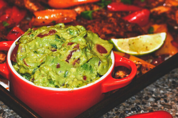 Image of guacamole in a red serving bowl next to a lime and sheet pan chicken fajitas