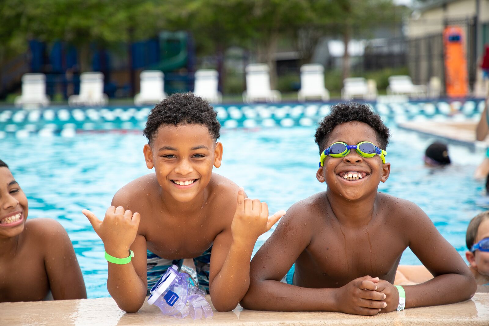 Three boys in pool smiling with one wearing swim googles and the other with his thumbs out two each child beside him