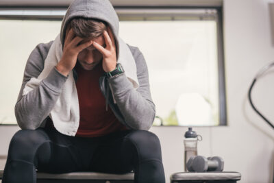 Strong sporty man sitting on gym bench with towel on face, suffering breakdown to overcome. Frustration sport concept.