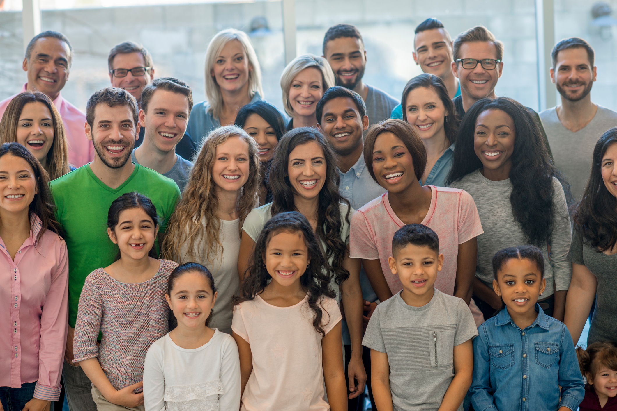 A multi-ethnic group of people are standing together in a room smiling and laughing. This is a diverse group of adults and children, with young children and seniors all together.