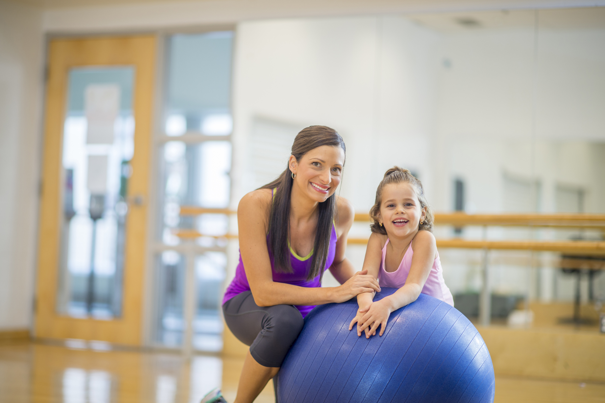 A mother and daughter are wearing athletic clothing and are happily together at the gym. They are smiling and looking at the camera.