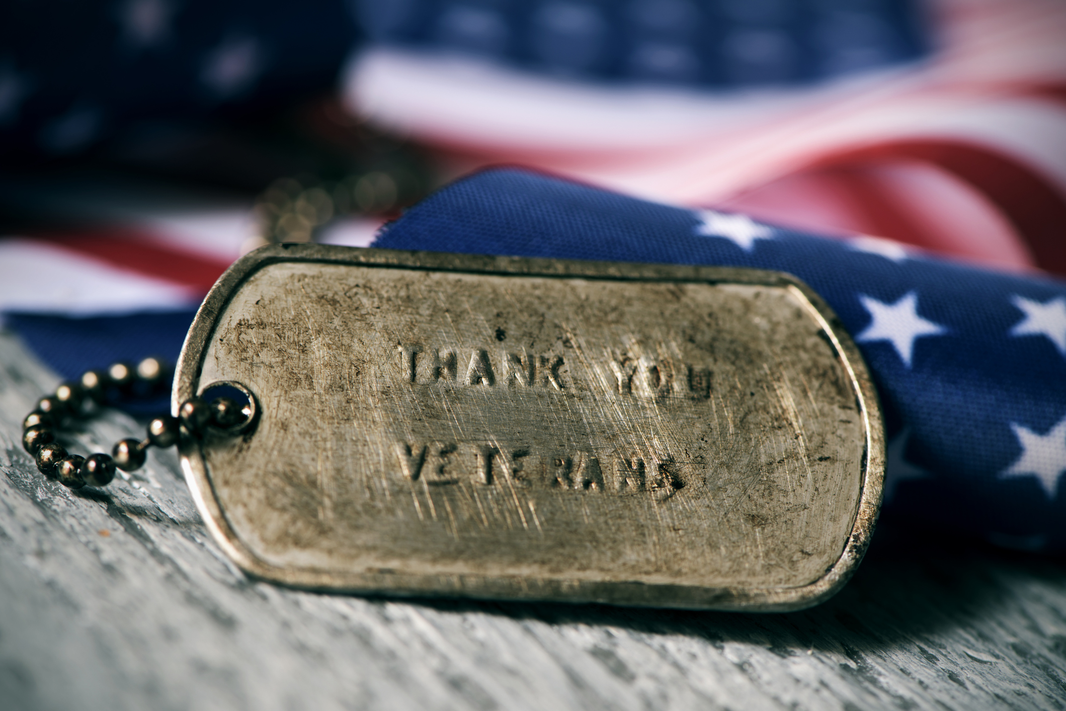 closeup of a rusty dog tag with the text thank you veterans engraved in it, next to a flag of the United States, on a rustic wooden surface