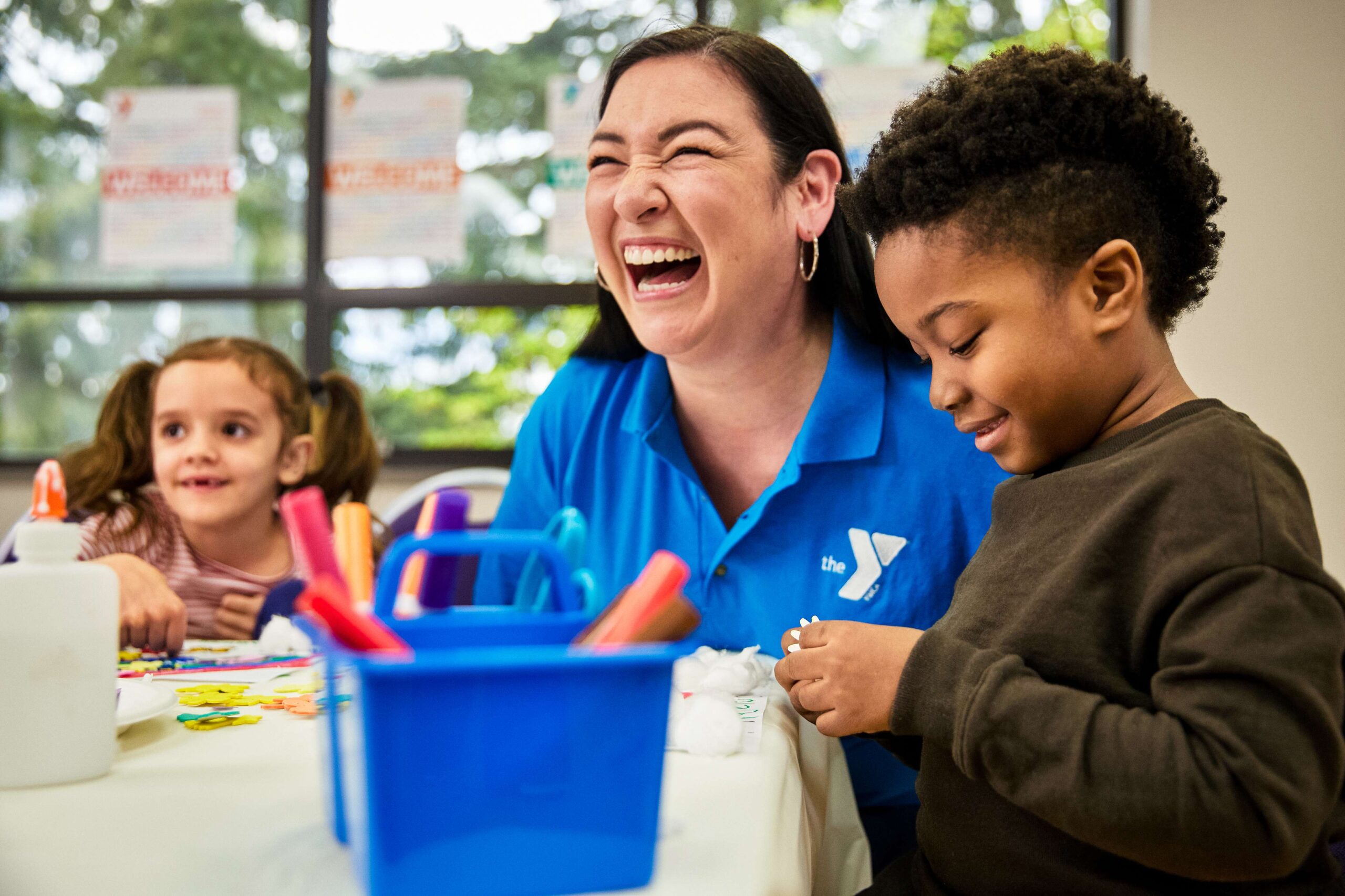 Woman smiling happily while wearing YMCA staff shirt while sitting next to two smiling children