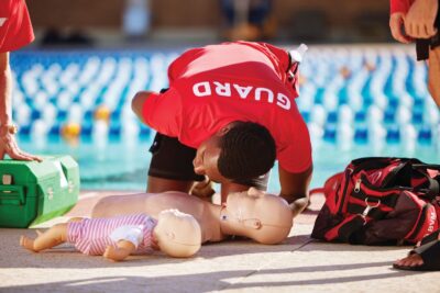 Lifeguard putting ear to a CPR doll on the pool deck beside the dool