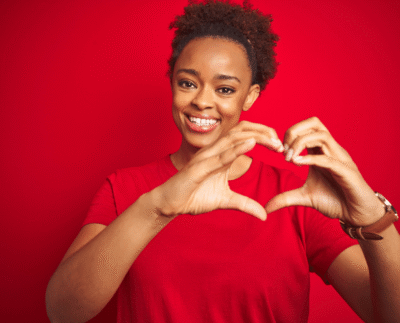 Woman smiling, holding hands together in a heart shape, with a red background, celebrating heart health.