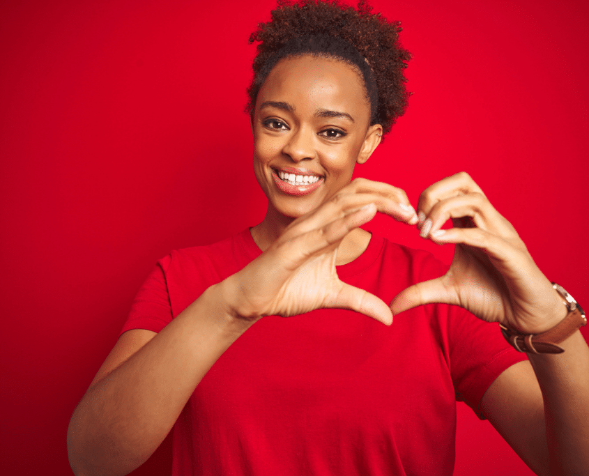 Woman smiling, holding hands together in a heart shape, with a red background, celebrating heart health.