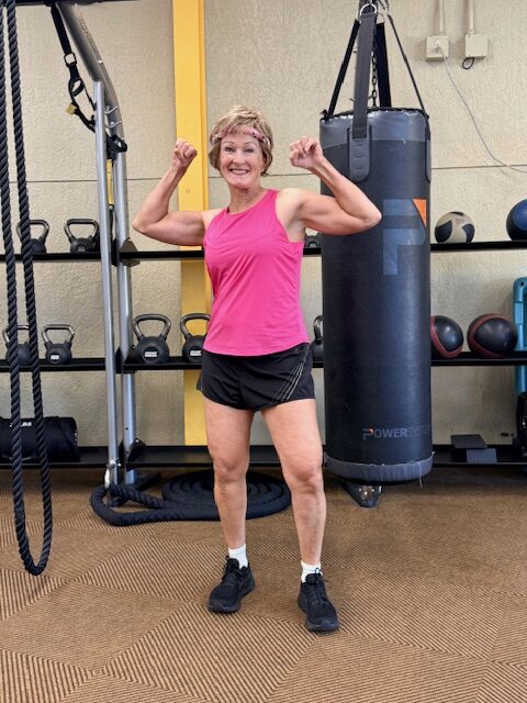 Woman in a pink tank top and black shorts flexing her muscles in a gym next to a punching bag and kettlebells.