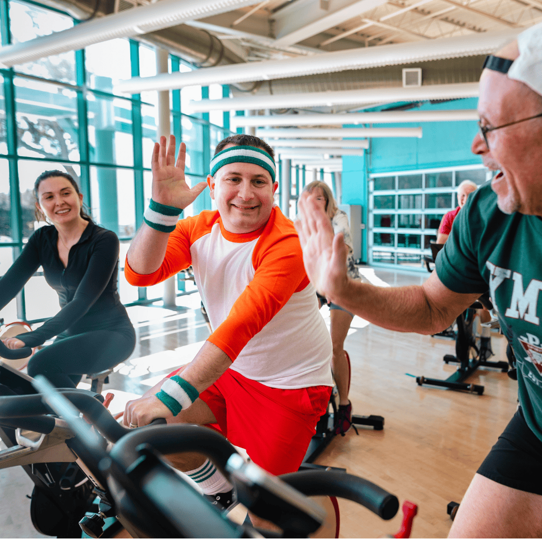 Three people in a cycling class at Roper YMCA, with one participant high-fiving another during the workout