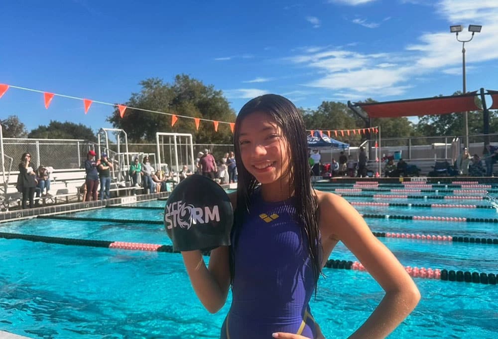 Girl holding up the Lake Nona YMCA Storm swim team cap in front of a pool