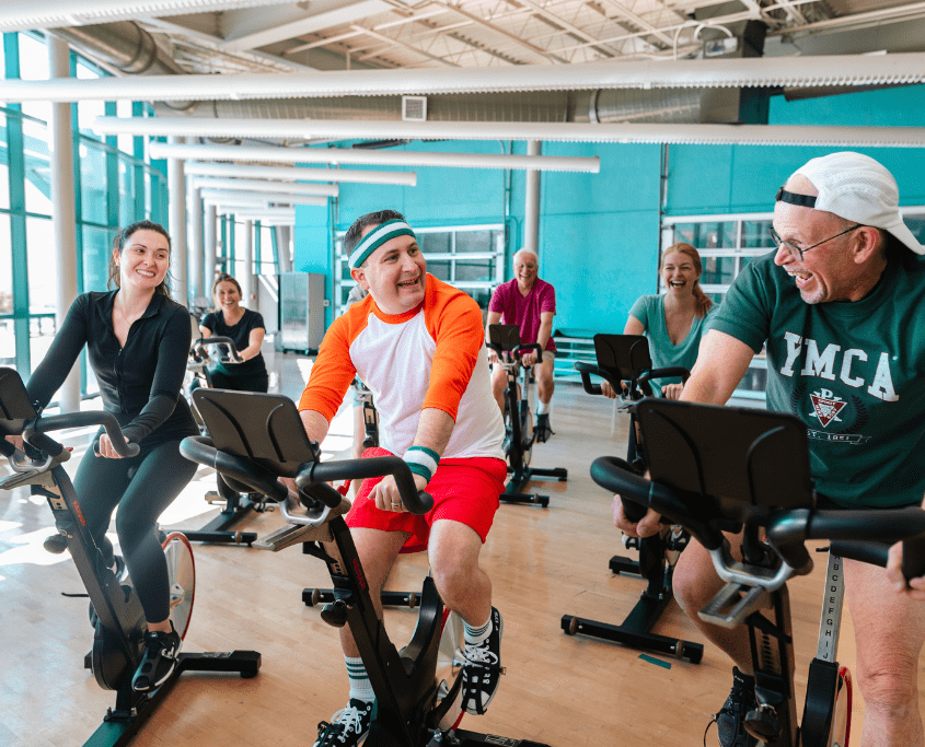 Three YMCA members participating in a group cycling class, riding stationary bikes in a fitness studio.
