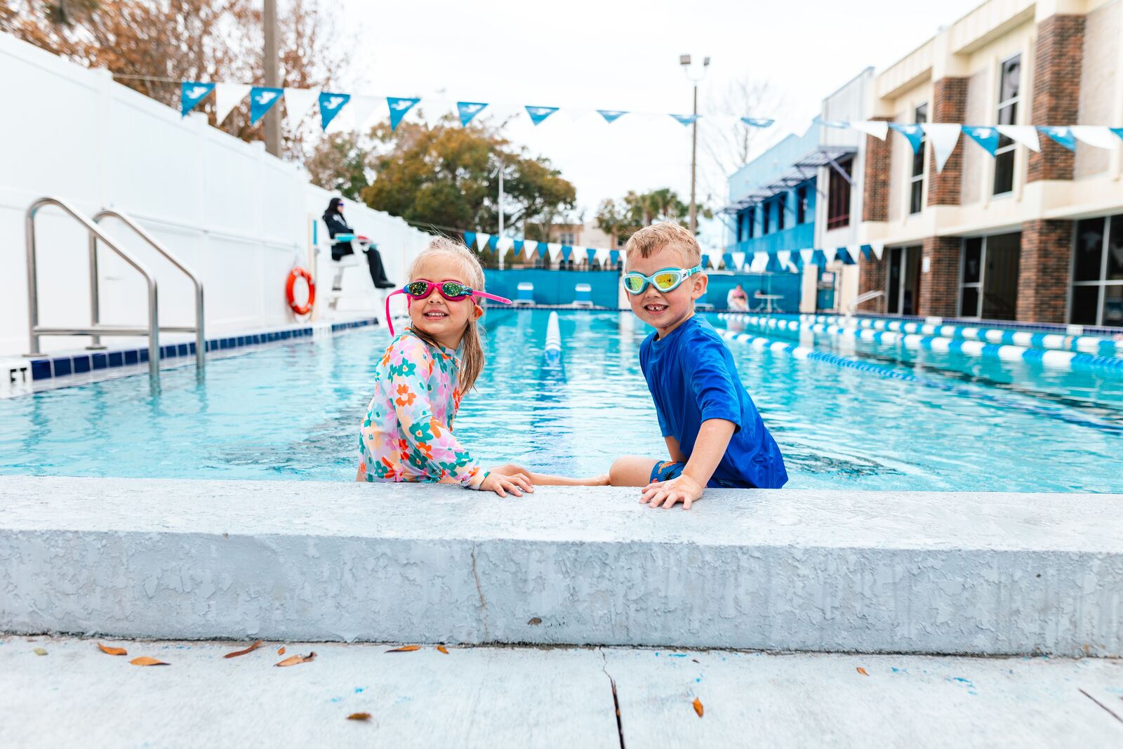 Two smiling children wearing swim goggles stand poolside at a YMCA swimming pool.