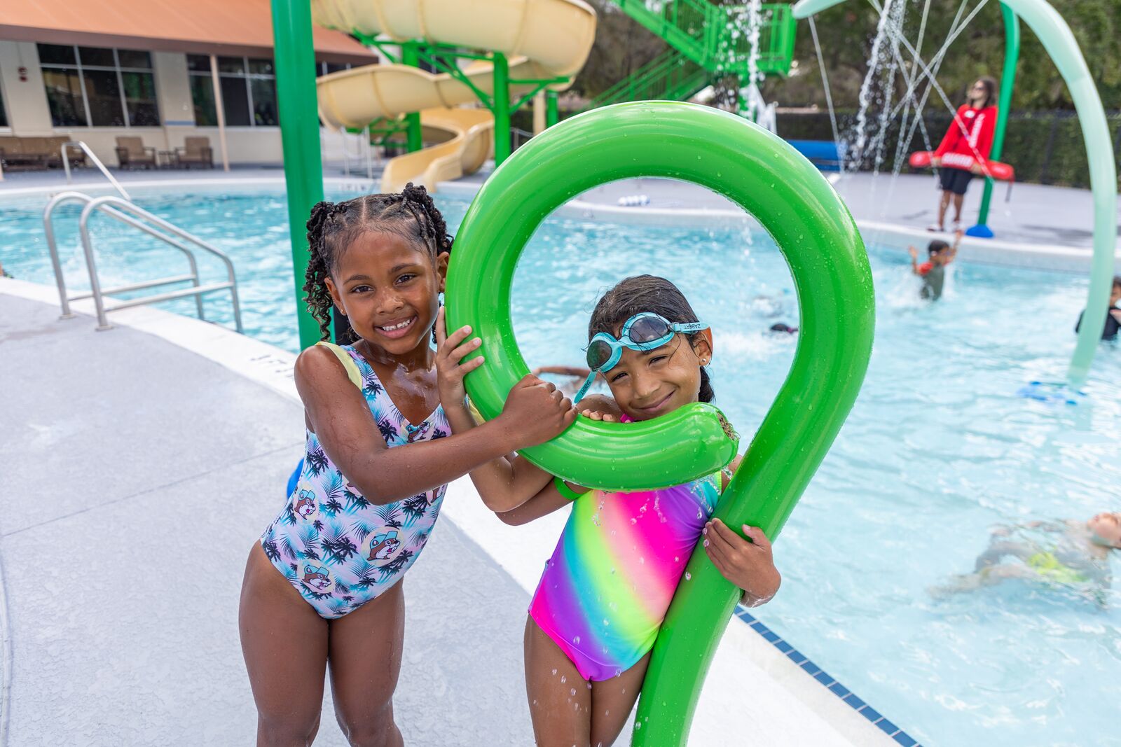 Two young girls smile and play in a YMCA splash pad water feature on a sunny day.